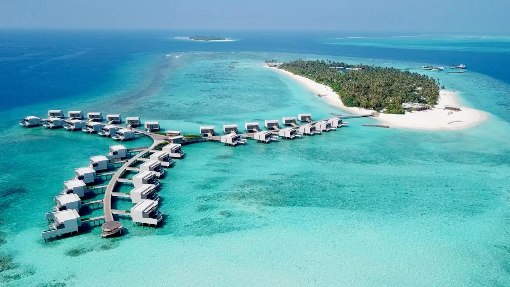 Aerial view of a luxury Maldivian resort showing overwater villas extending from a palm-covered island into turquoise lagoon waters