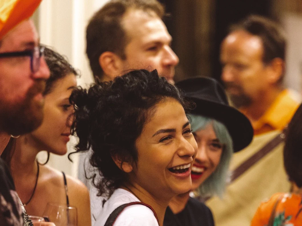 A diverse group of people, including a woman with curly hair laughing, socialize at an indoor event, holding drinks and wearing wristbands.