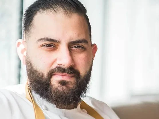 Portrait of a bearded chef in a white chef's coat and denim apron, seated at a restaurant table with a watch on his wrist.