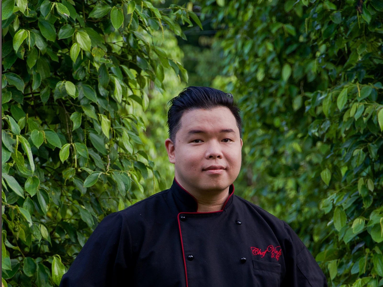 A person in professional black and red chef's attire stands in front of dense green climbing vines or ivy.