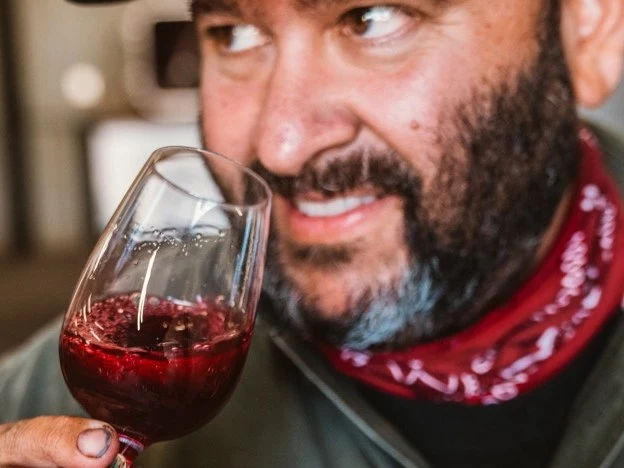 A bearded man in a green jacket and red bandana holds a glass of red wine, examining it closely in a dimly lit setting.