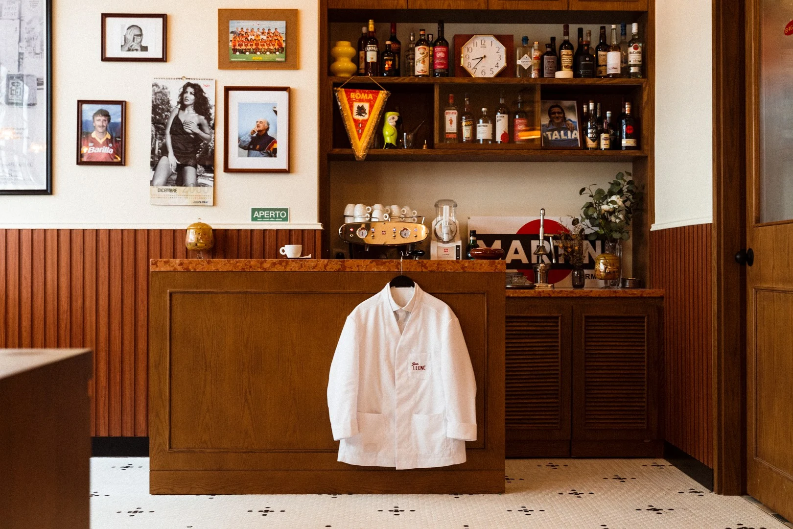 Interior of an Italian-themed bar showing a wooden bar counter with espresso machine, shelving with liquor bottles, framed photographs on walls, and a