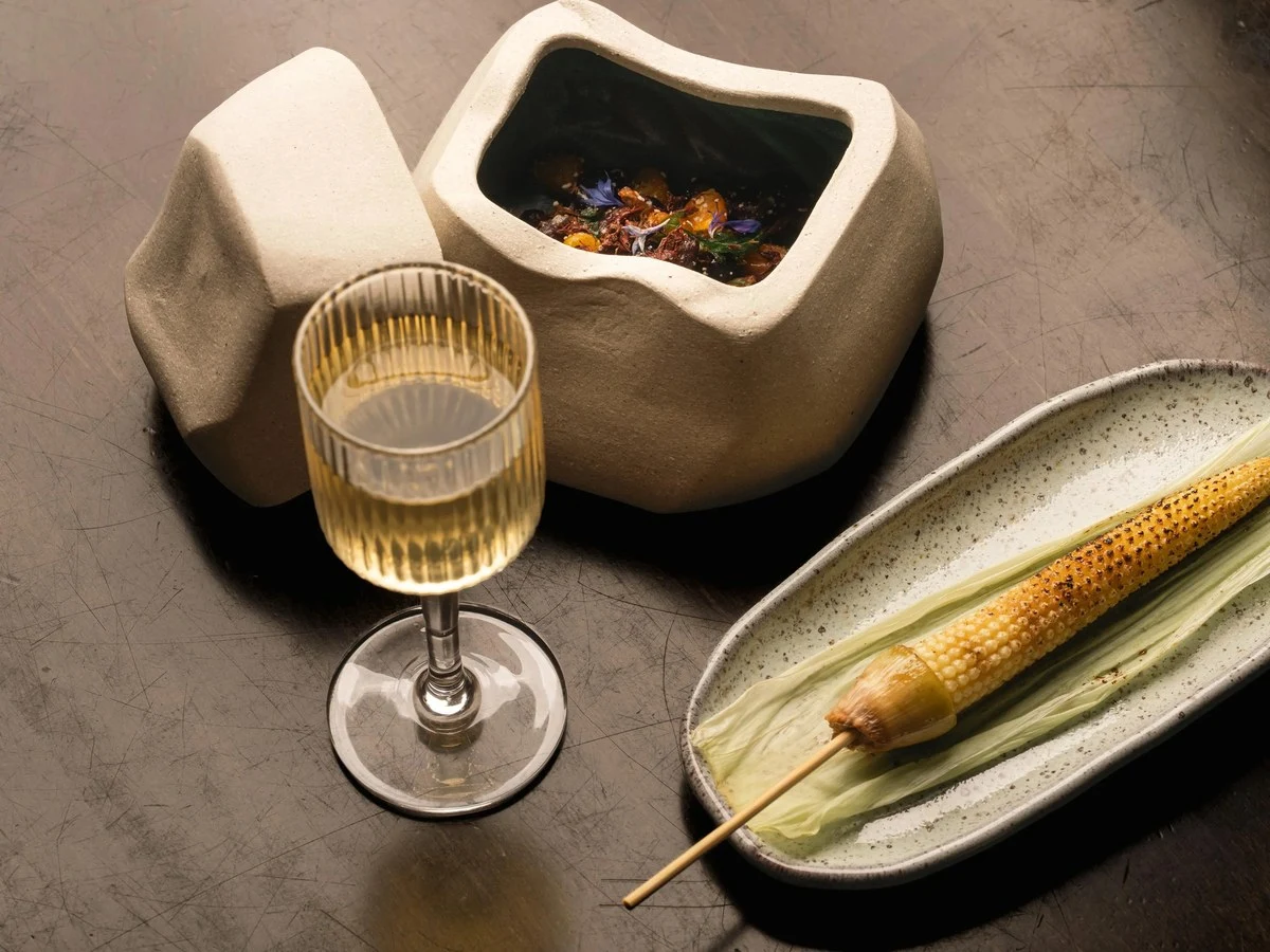Editorial food photography showing an ear of grilled corn on the husk, a glass of white wine, and a sculptural ceramic vessel containing what appears 