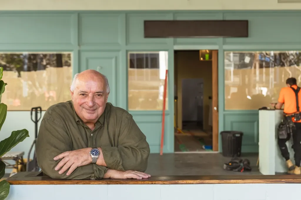 A bald man wearing an olive button-up shirt and watch stands at a wooden counter inside a contemporary retail space with turquoise paneling and large windows.