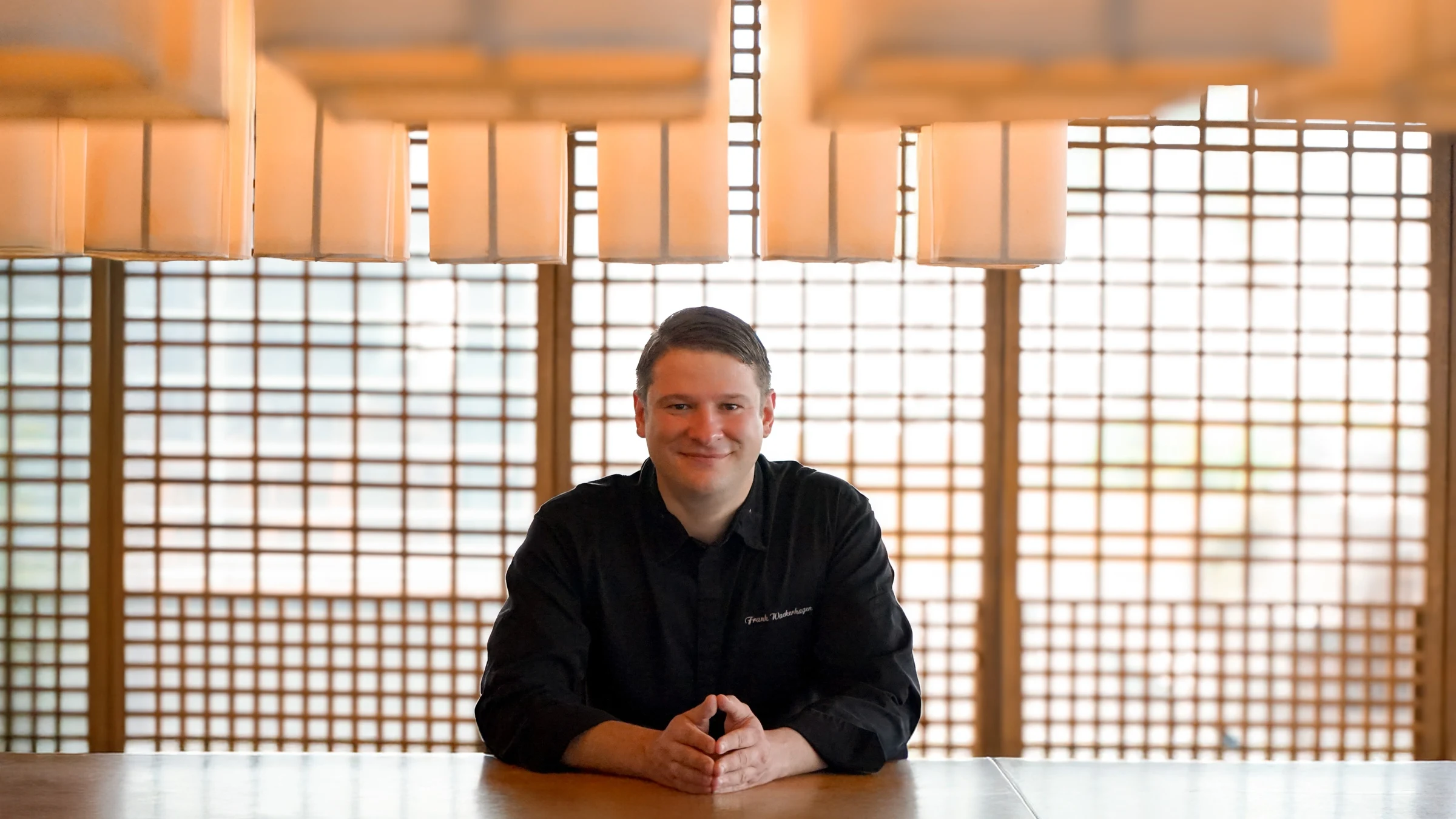Professional portrait of a chef in black chef's coat seated at a table in a dining space with Japanese-style shoji screens and warm pendant lighting; 