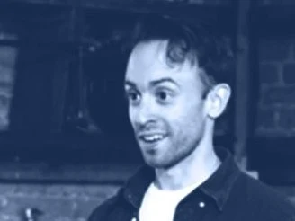 A man wearing a dark jacket over a white shirt, smiling and looking at the camera in an indoor setting.