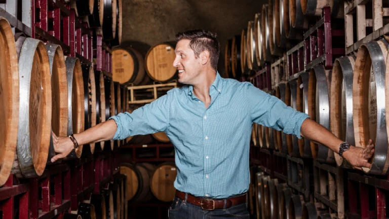A man in a light blue button-down shirt stands with arms outstretched in a wine barrel cellar