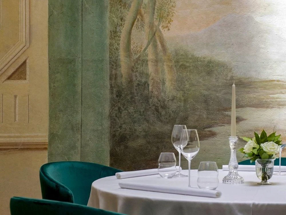 Fine dining table setting with white tablecloth, place settings, wine glasses, white candle, and fresh flowers, with teal upholstered chairs and a painted landscape mural on the wall.