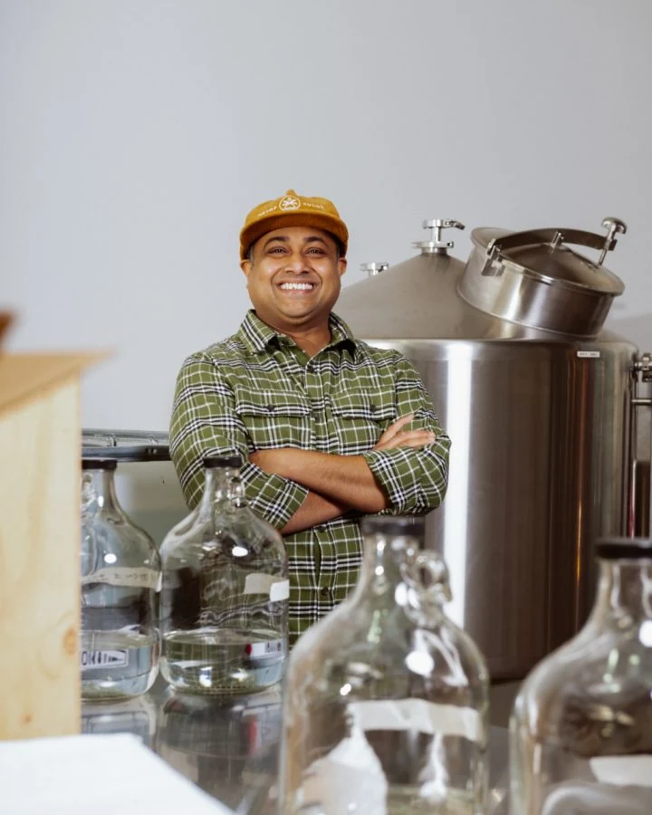 A smiling man in a plaid shirt and yellow cap stands with arms crossed in front of distillery equipment (stainless steel tank) and empty glass bottles