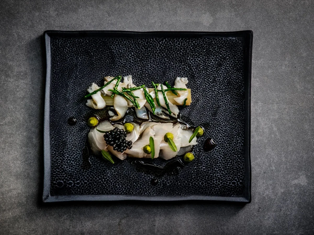 A plated FYN dish: delicate seafood with caviar, green herbs, and small spherical elements in a dark broth.