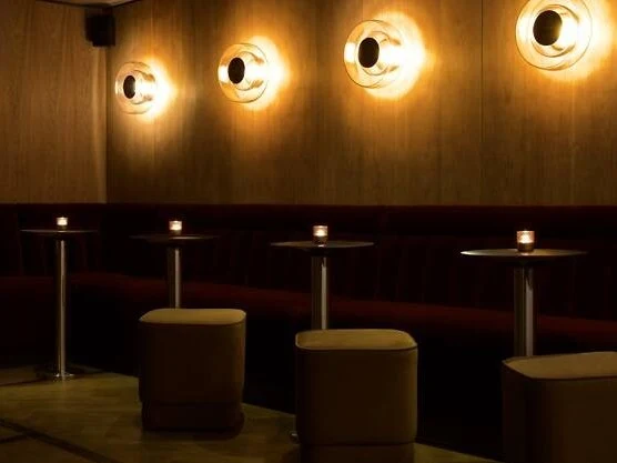 A dimly lit bar interior with four round illuminated wall fixtures above dark booth seating and cylindrical ottomans on a light floor.