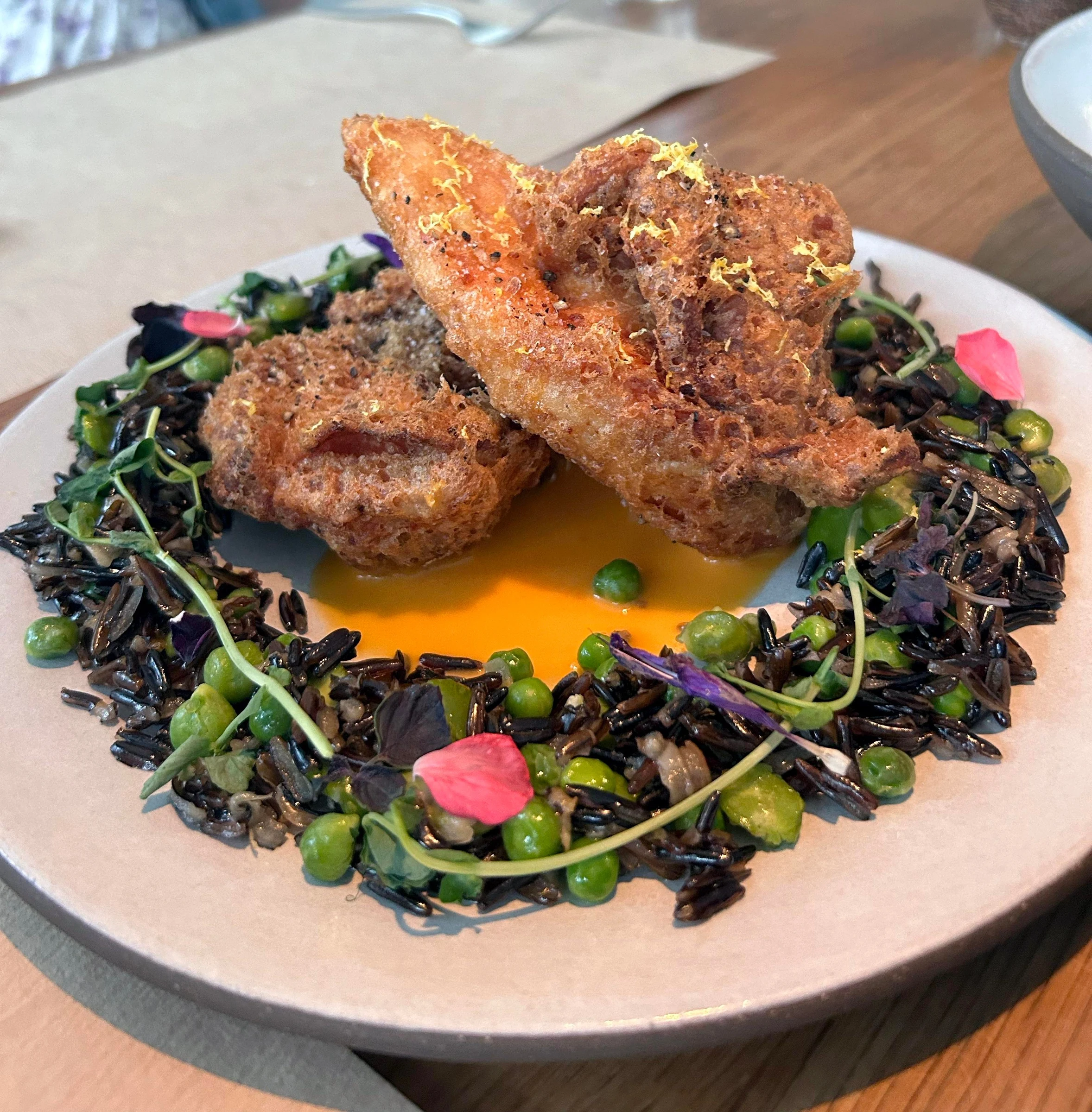 A plated dish featuring breaded, fried protein (likely shrimp) atop black rice with edamame and microgreens, with an orange sauce pool.