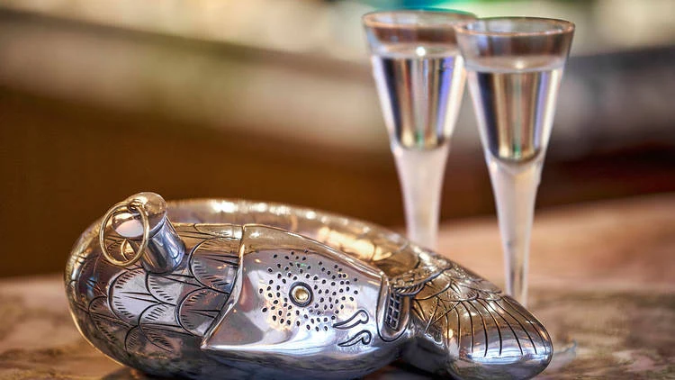 A decorative silver fish-shaped dish with detailed scales and fins sits on a wooden surface next to two tall champagne glasses filled with clear liquid.