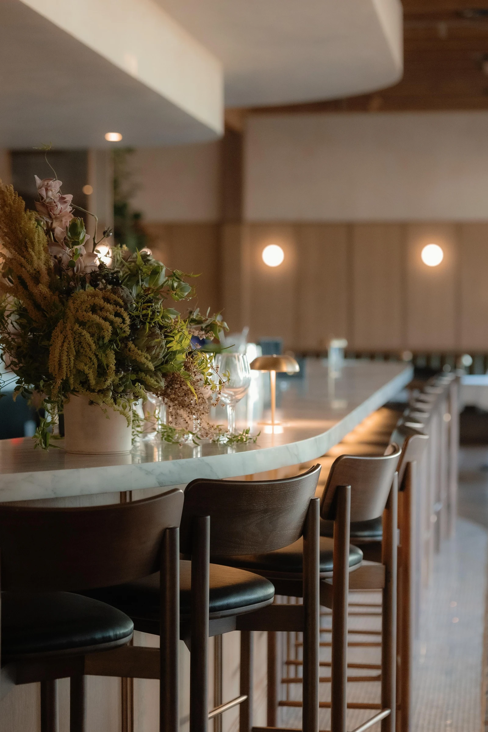 A sleek white marble bar with wooden bar stools, floral arrangement, and modern pendant lighting in an upscale restaurant interior