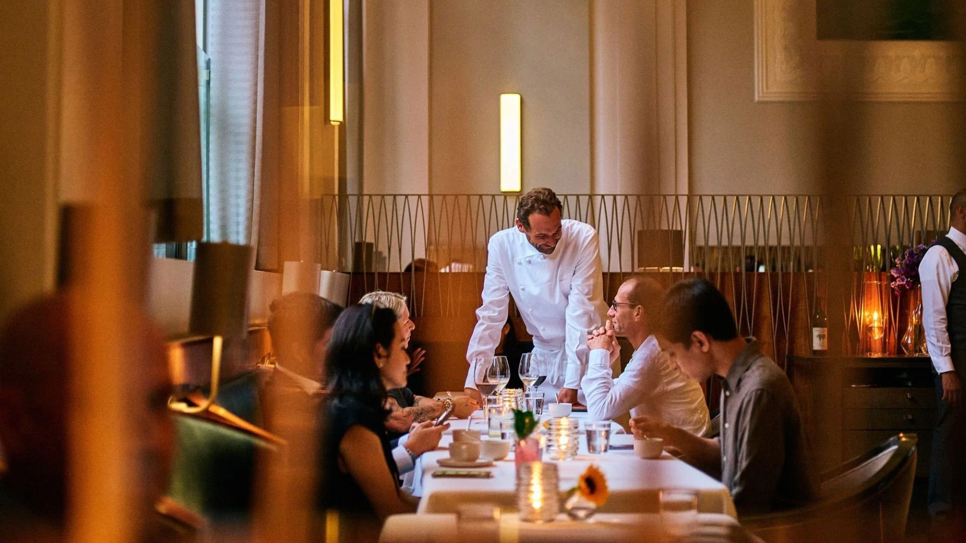 A male chef in a white chef's coat leans toward a table of four diners in an upscale restaurant with warm lighting and modern decor.