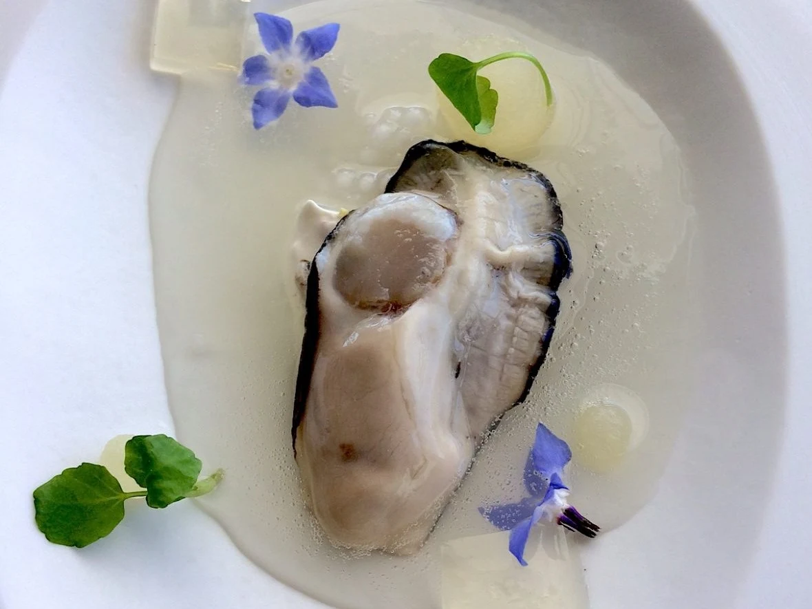 Oyster on a plate with edible flowers and a clear liquid.
