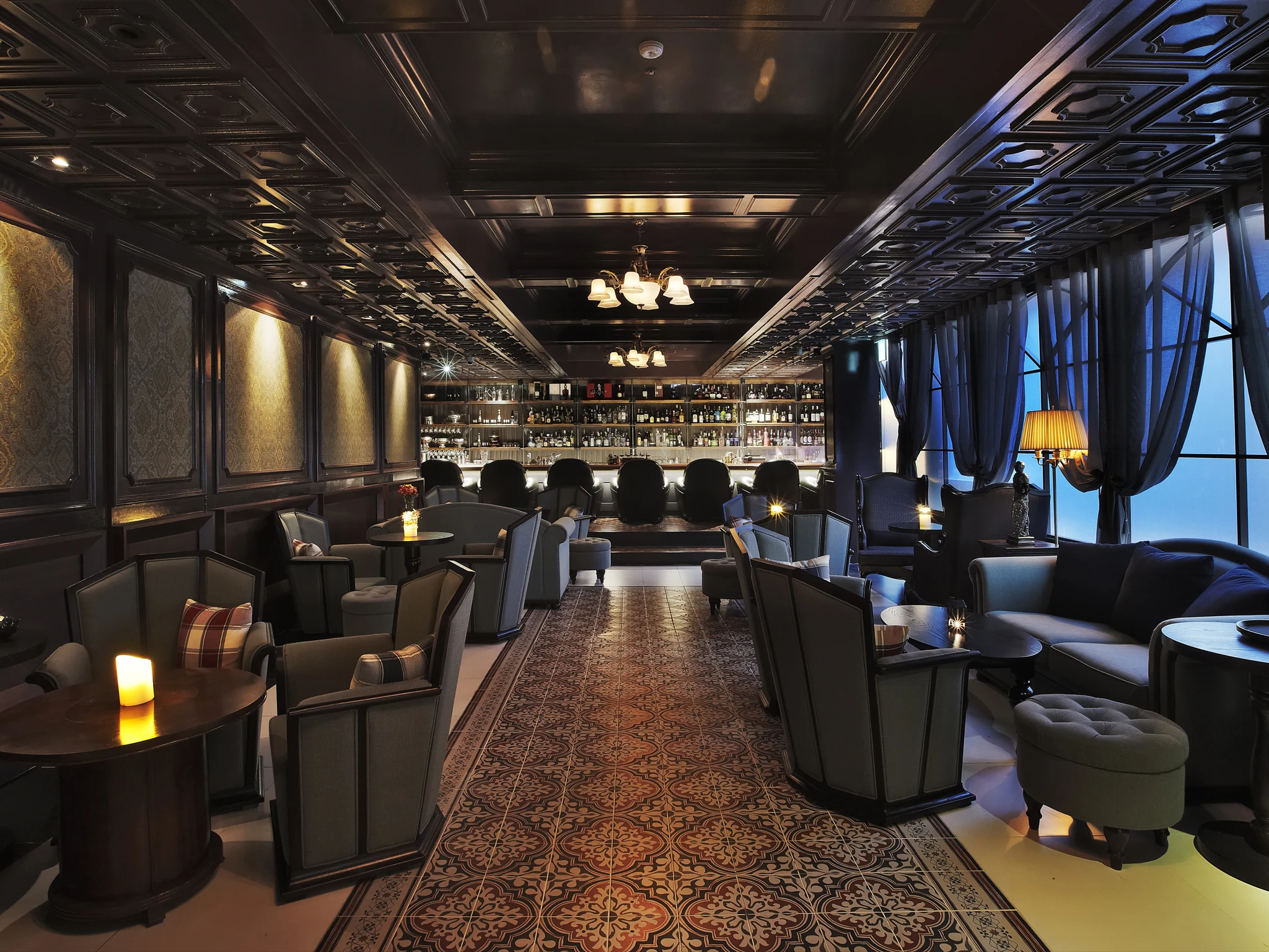 An upscale lounge bar with dark coffered ceilings, plush seating, a patterned floor runner, and a brightly lit bar counter.