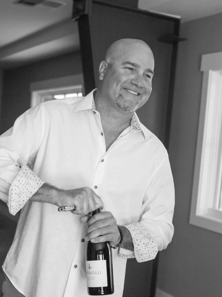 A professional portrait of a bald man in a white dress shirt holding a wine bottle, photographed indoors
