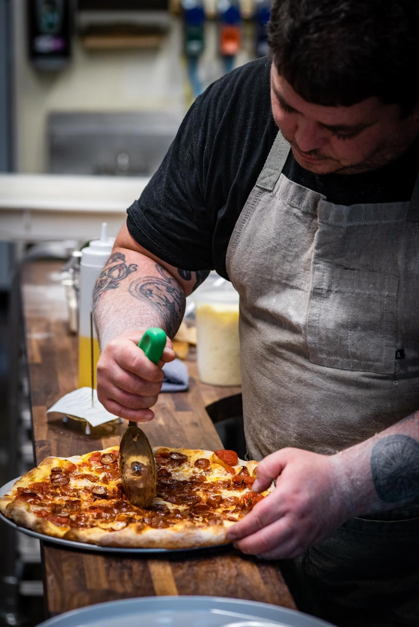 A man wearing a gray apron and black shirt cuts a pepperoni pizza with a green-handled pizza cutter on a wooden counter.