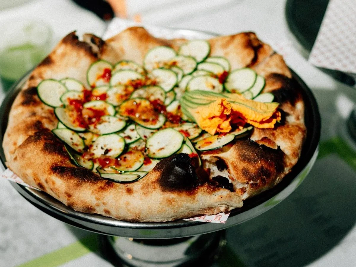 A finished pizza with thinly sliced zucchini ribbons, edible flowers, and chili flakes on a charred, high-hydration crust, served on a black pan.