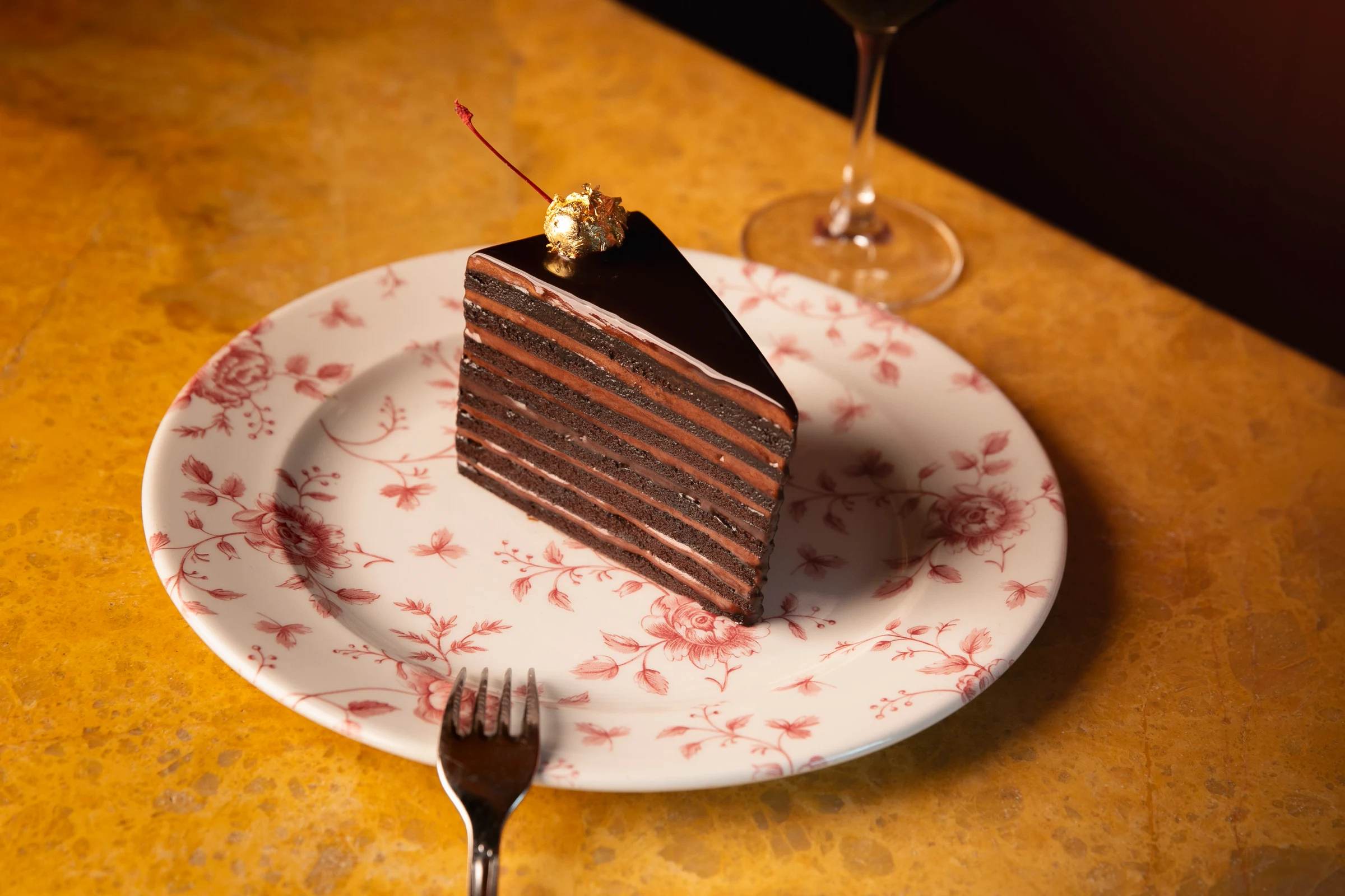A slice of layered chocolate cake with dark ganache coating and gold leaf decoration on a white plate with red floral pattern, with a fork and glass visible.