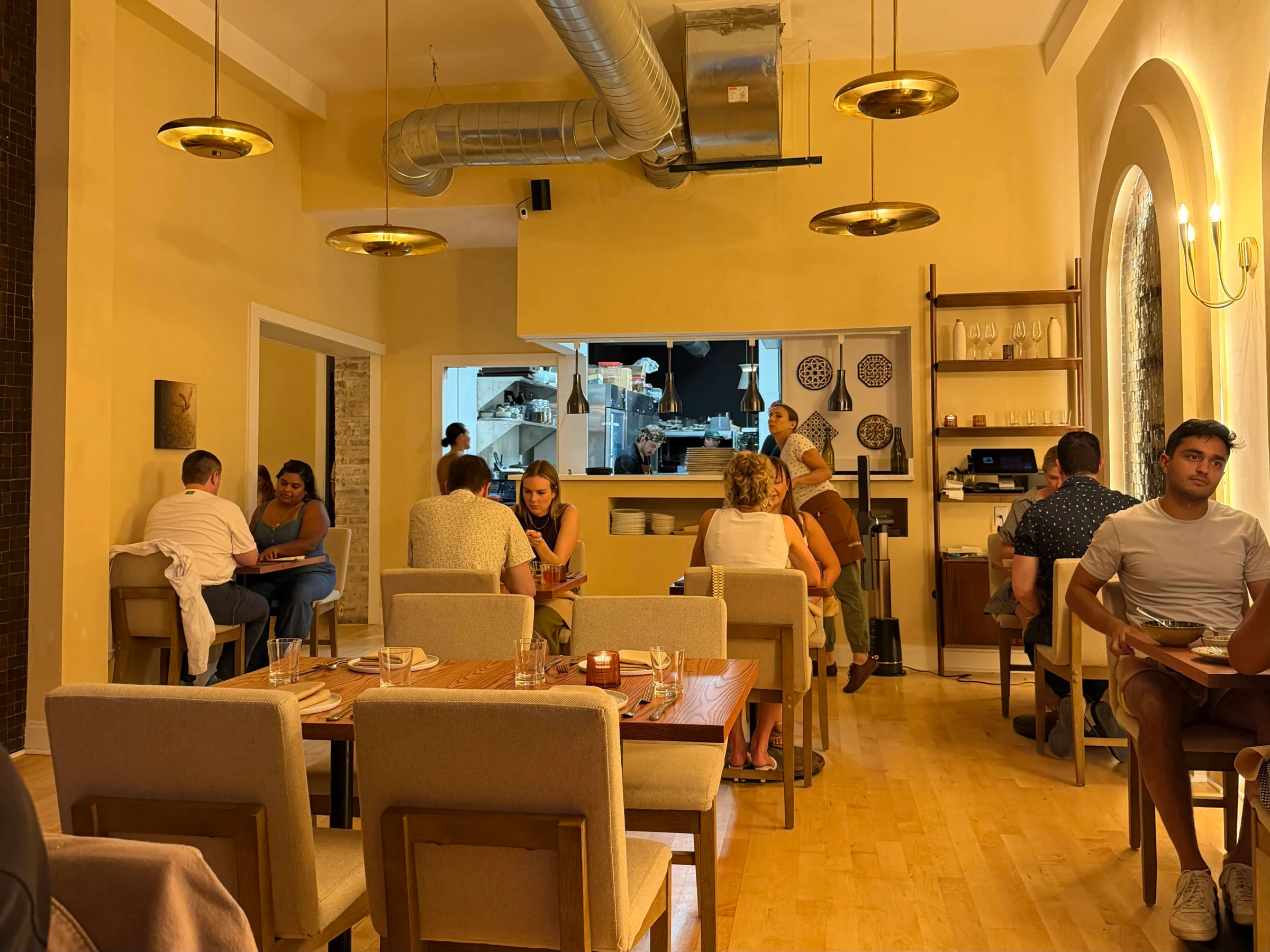 A modern restaurant dining room with warm lighting, beige upholstered chairs, wooden tables set with glassware and candles, exposed ductwork, pendant 