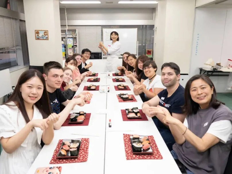 A group of students and instructors at Kudan Japanese Institute, seated around a long table with plates of sushi, smiling for a photo.