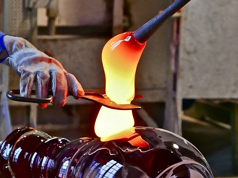 A glassblower's gloved hands use metal jacks to shape glowing molten glass on a blowpipe, with a finished red glass vessel below.