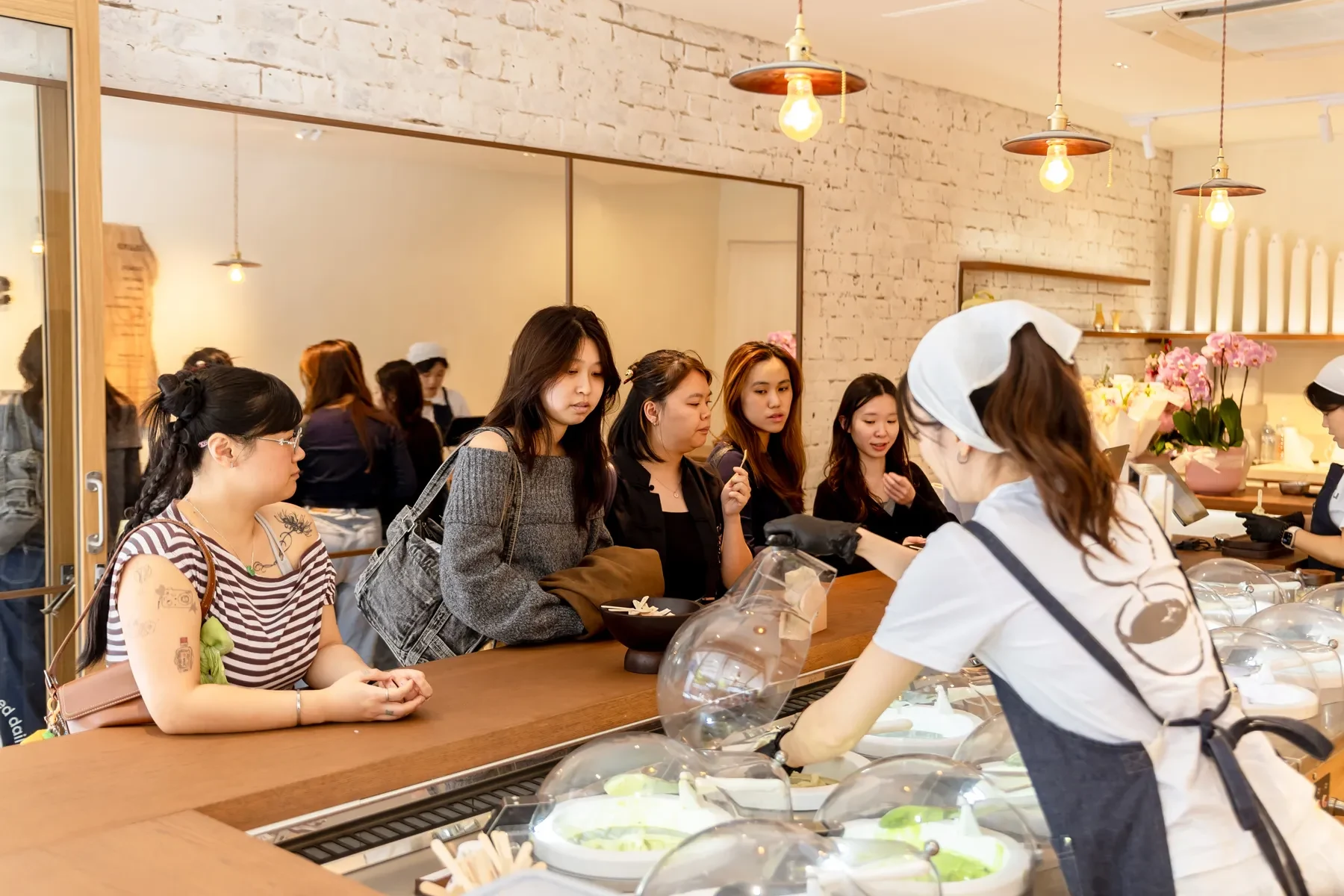 Counter dining scene at a French restaurant showing a staff member in white uniform and headband demonstrating or serving to seated customers at a woo