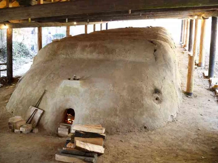 A large, light brown, mud-built climbing kiln with a small arched opening at its base, where a fire glows, is sheltered under a wooden roof structure.