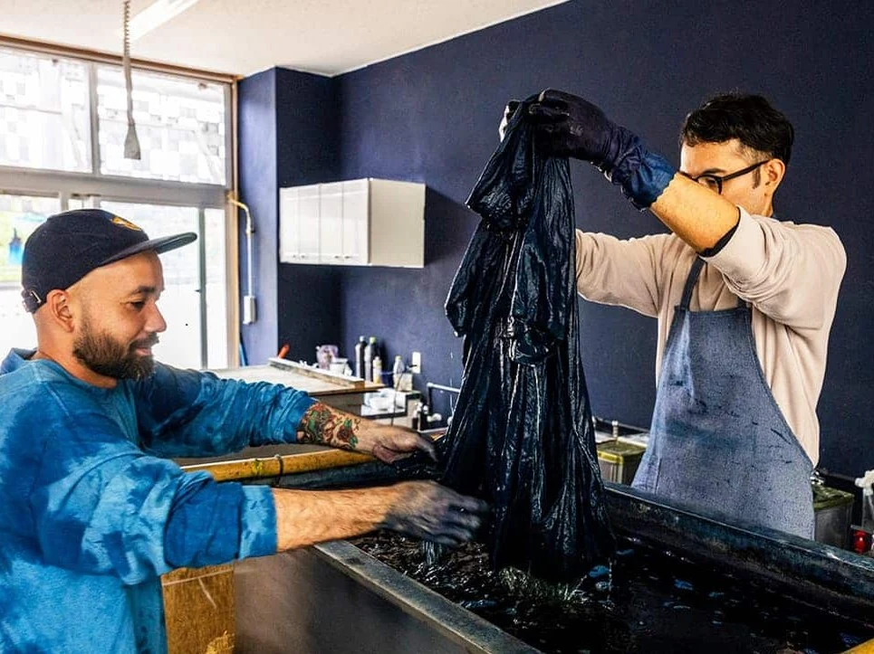Two craftspeople, one with a beard and a blue tie-dye shirt, the other in an apron and glasses, work at an indigo dye vat, handling dark-dyed fabric