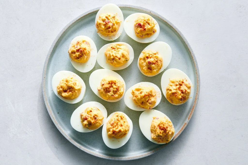 Twelve deviled eggs arranged on a pale blue ceramic plate, each topped with a yellow-orange filling garnished with red spice flakes; the source page e