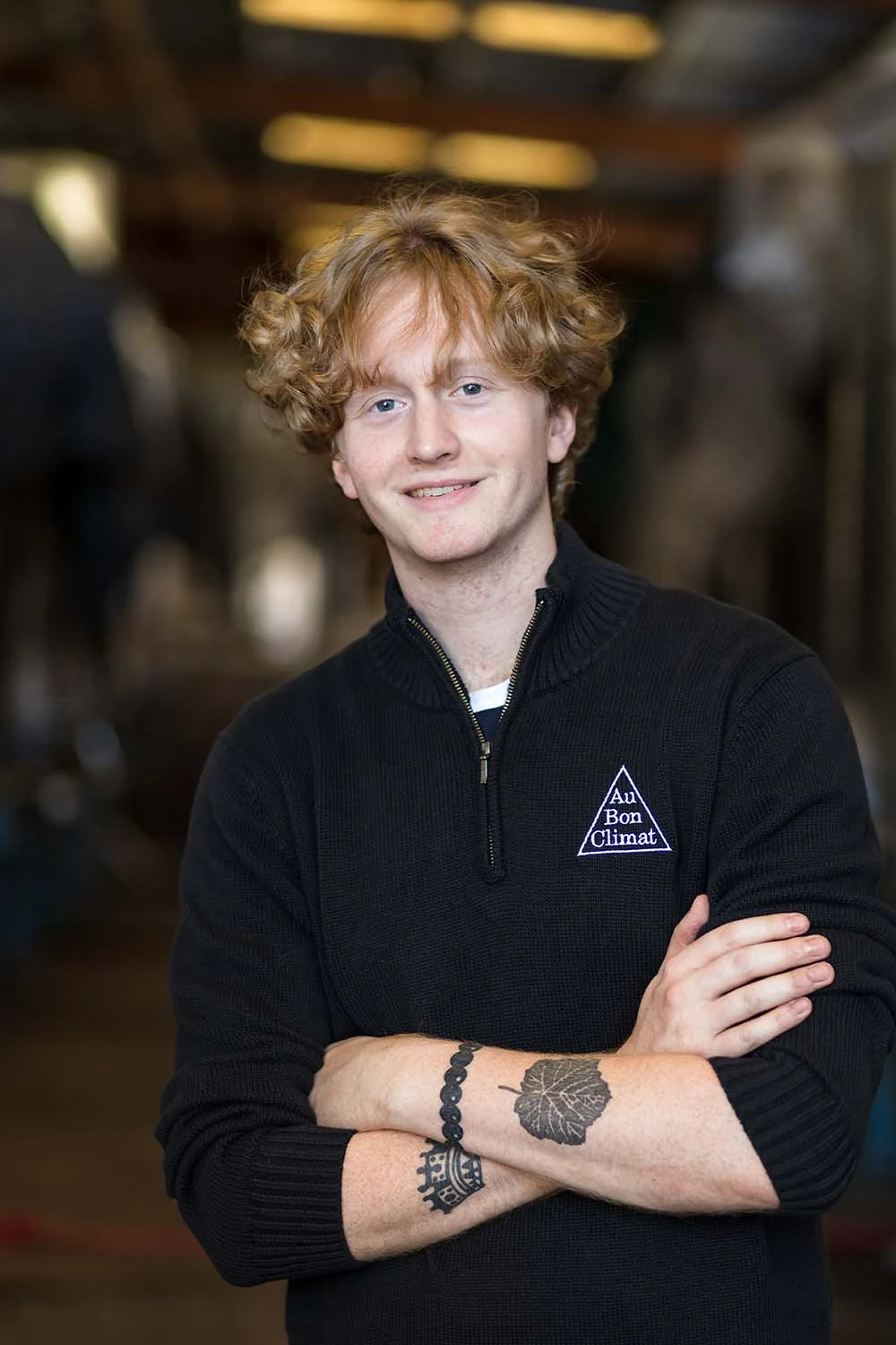 A young man with curly blonde hair wearing a black quarter-zip sweater with 'Au Bon Climat' logo, arms crossed, standing in what appears to be an indu
