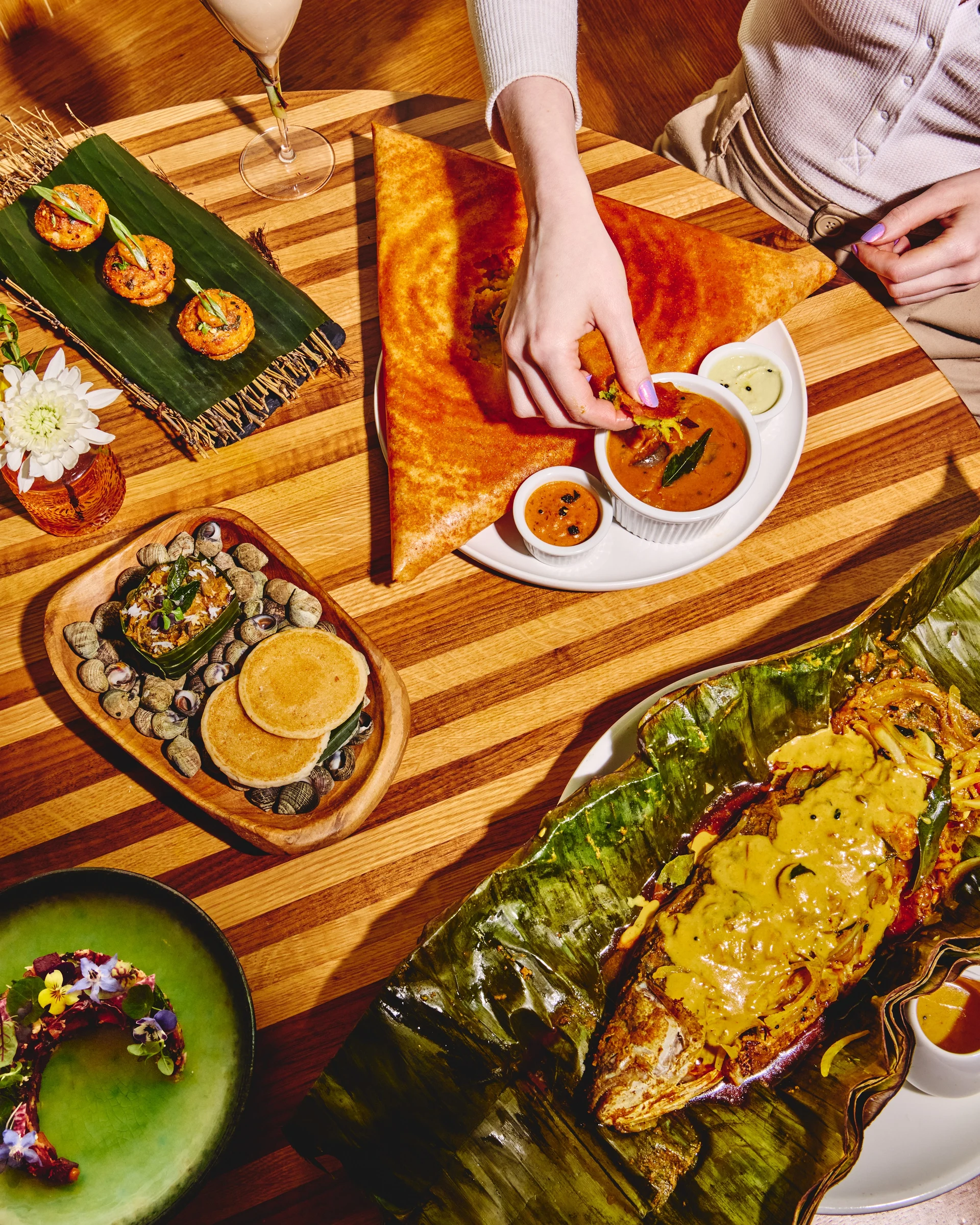 An overhead shot of an Indian restaurant table spread featuring multiple dishes including a large dosa, small bites on banana leaf, grilled seafood, a