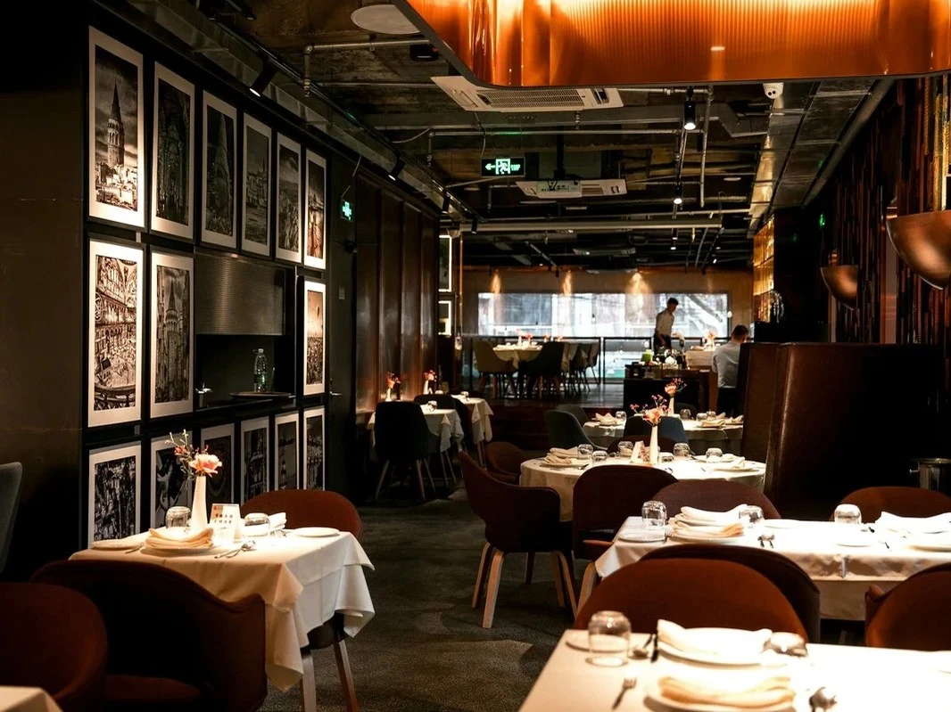An upscale restaurant dining room with warm lighting, dark walls featuring framed black-and-white photographs, white-tablecloth tables with brown.