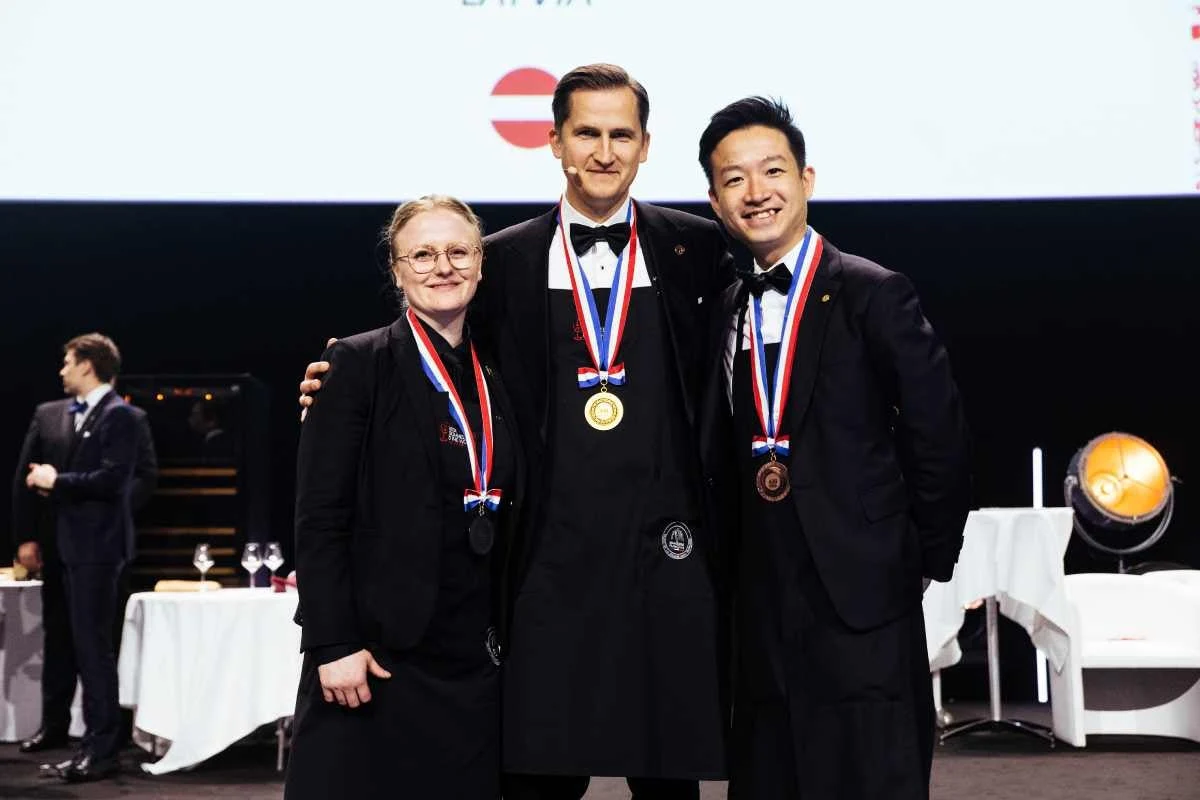Three sommeliers in formal attire wearing medals on ribbons stand on a competition stage with white-clothed tables visible