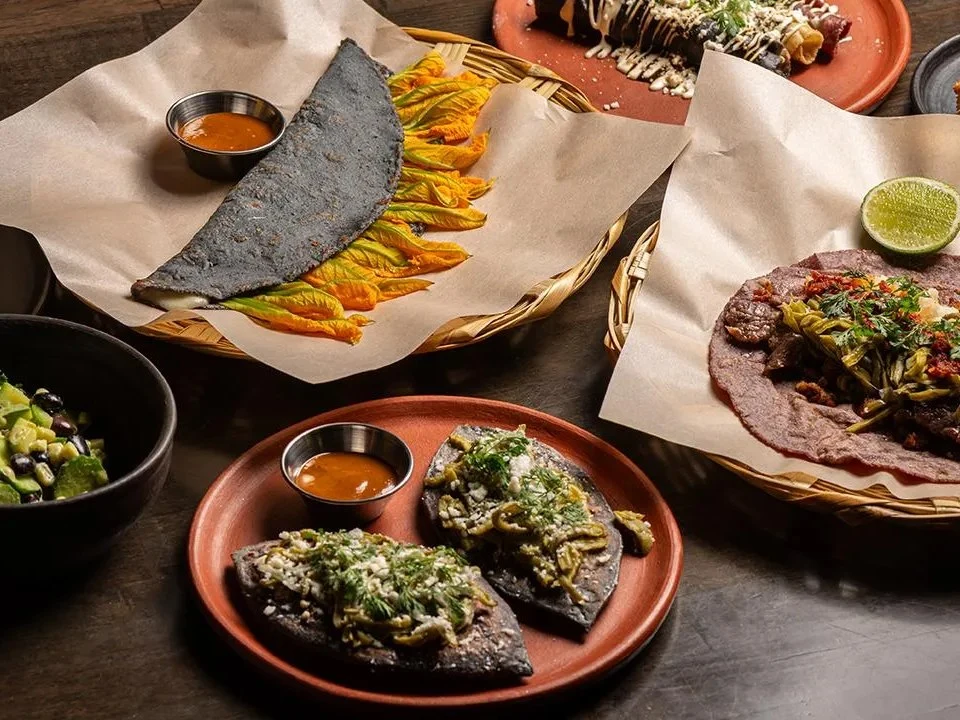 A spread of Mexican dishes featuring blue and purple corn tortillas with various toppings, presented on terracotta plates and woven baskets; the sourc