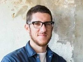 Mitch Orr, the chef who departed The Palomar, poses in a navy jacket against a weathered wall.