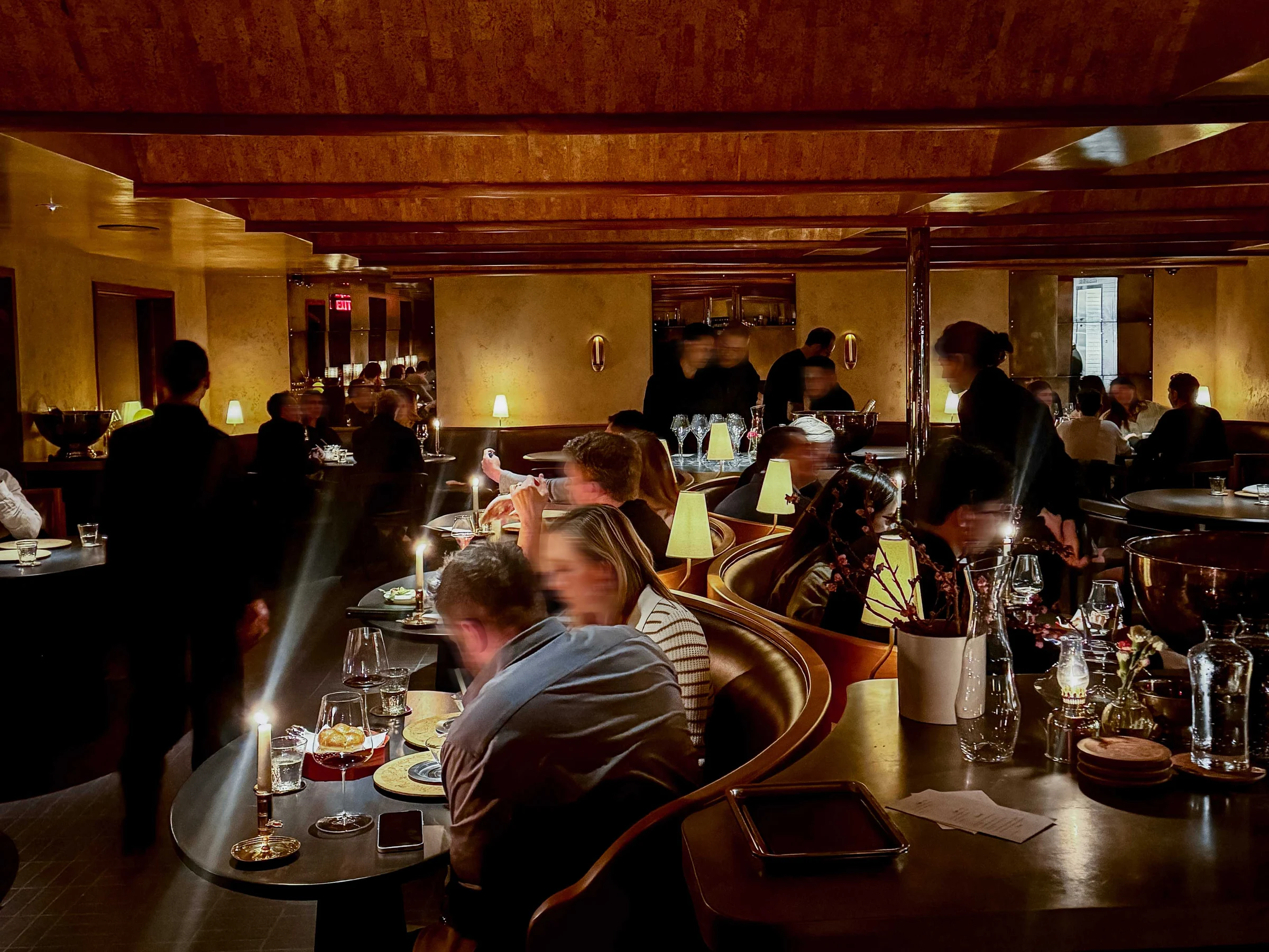 An upscale restaurant dining room with warm golden walls, curved banquettes, intimate table lamps, and diners at small tables.