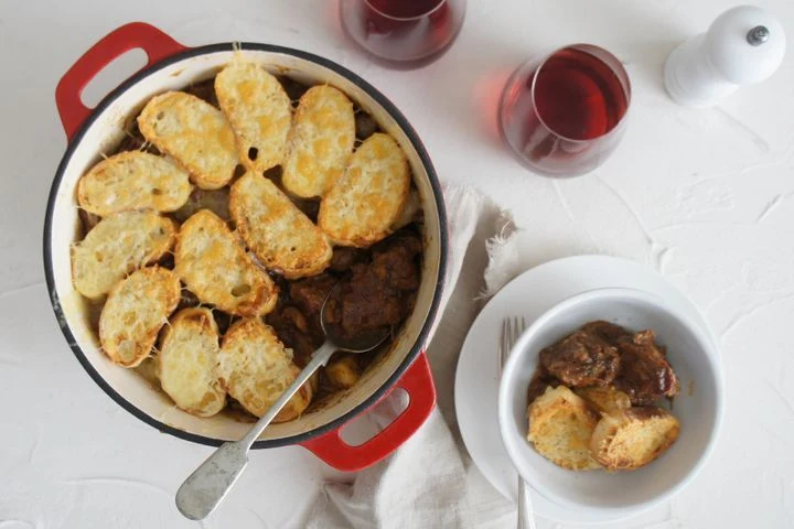 A casserole dish containing braised meat topped with toasted bread slices, served with red wine