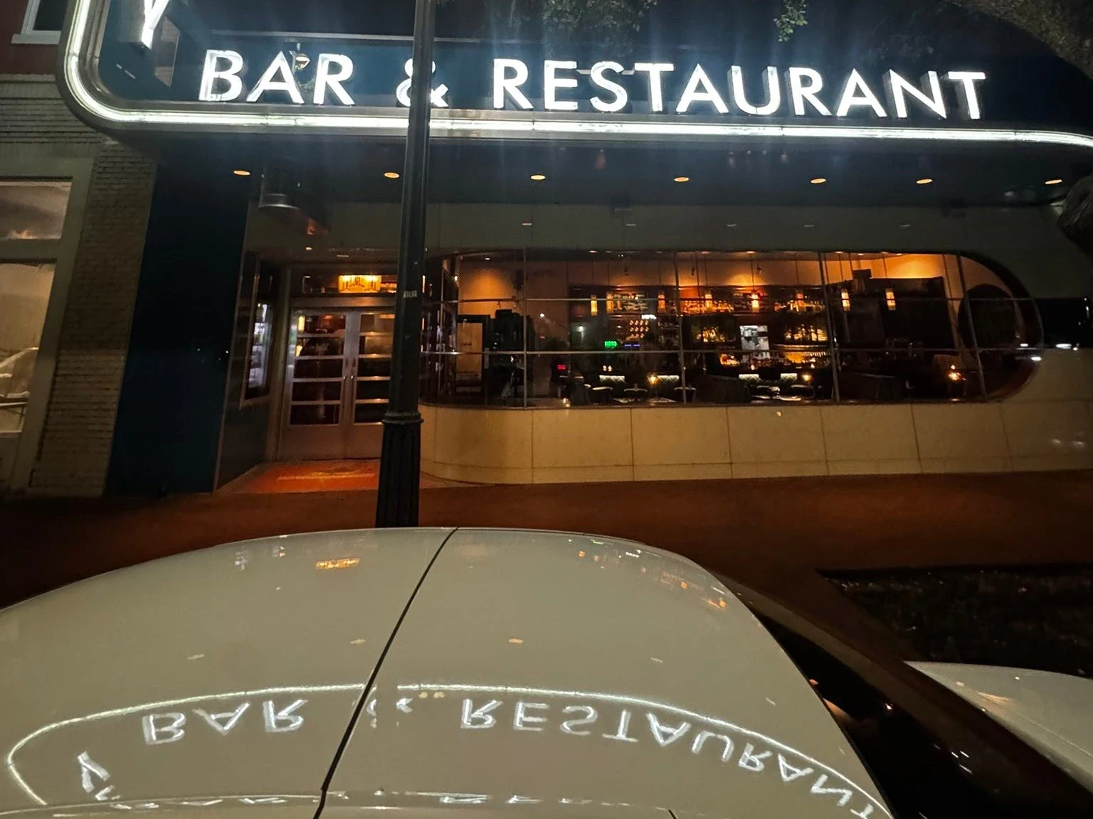 Illuminated storefront with 'BAR & RESTAURANT' sign at night, with a street lamp in the foreground and a car hood visible at the bottom of the frame.