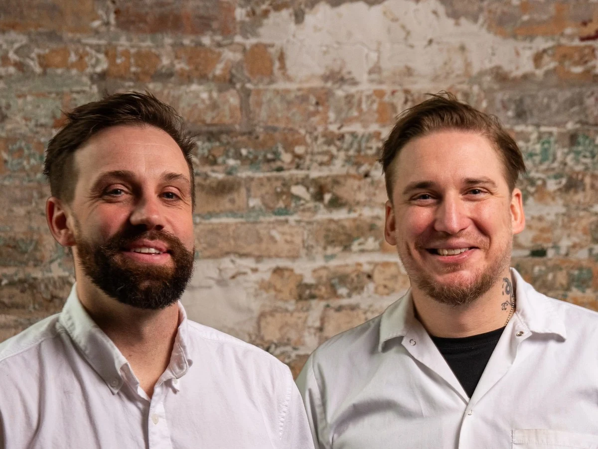 Two men, Larry Feldmeier and Noah Sandoval, smile at the camera while standing in front of an exposed brick wall.