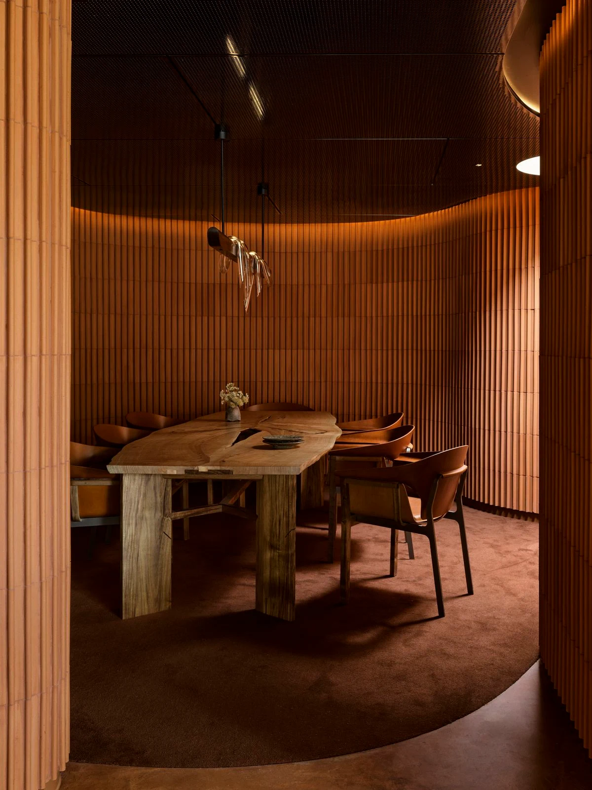 A contemporary fine-dining room with vertical timber slat walls, warm indirect lighting, a live-edge wooden table with distinctive cross-base, leather