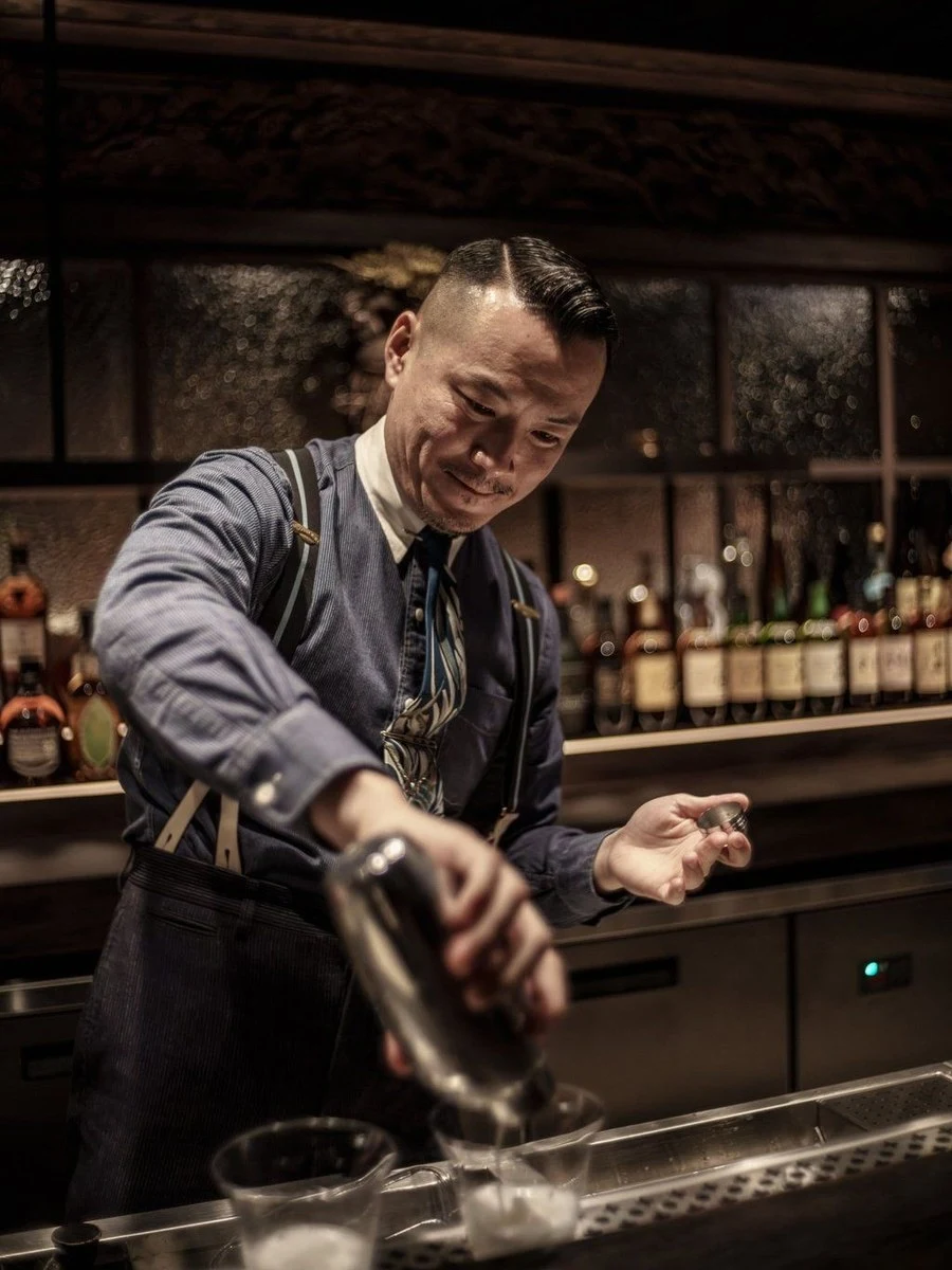 A male bartender in a blue pinstriped shirt and suspenders, with a neatly slicked-back haircut, pours liquid from a metal shaker into a glass.