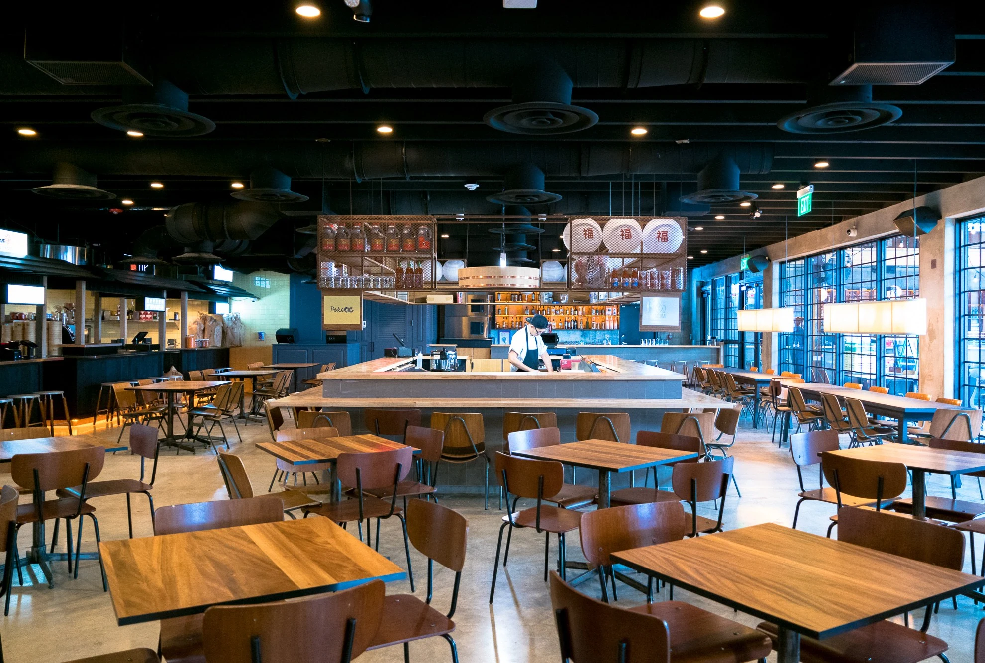 Modern food hall dining area with wooden tables and chairs, central service counter with staff, industrial ceiling, and Asian-themed signage visible o