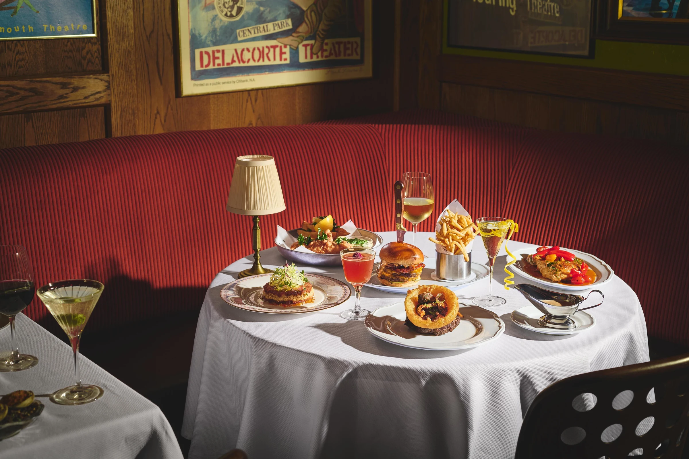 Fine dining table setup with multiple plated dishes including burgers and seafood, cocktails, wine glasses, and a table lamp against a red upholstered booth in a restaurant interior.