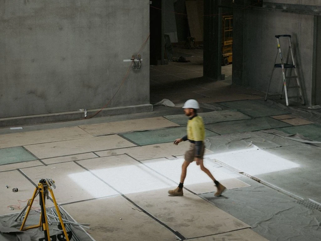 A construction worker in a hard hat and high-visibility vest walks across a large, tiled floor in an unfinished building, with concrete walls.