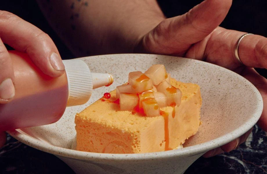 A dessert dish showing a rectangular parfait with diced pear topping and chilli oil being drizzled, held by hands over a speckled ceramic plate; sourc